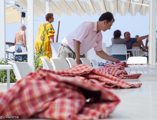 Un cameriere in camicia rosa e pantaloni beige è chino mentre stende delle tovaglie a quadri rossi e bianchi su una lunga tavola sotto un gazebo bianco. Sullo sfondo si intravedono alcuni clienti del ristorante all'aperto.