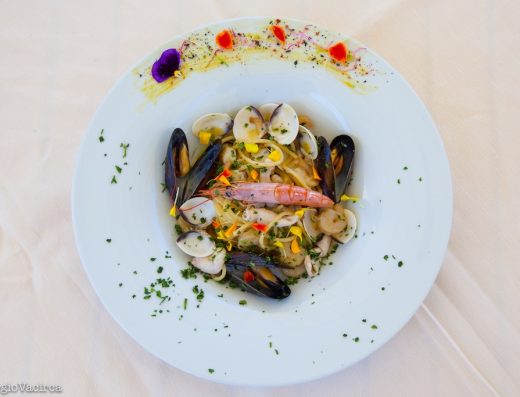 Inquadratura dall'alto di un piatto di spaghetti ai frutti di mare, decorato con cozze, vongole, calamari e un grande scampo al centro. Il bordo del piatto bianco è rifinito con una spennellata di zafferano, petali di fiori edibili colorati e prezzemolo tritato.